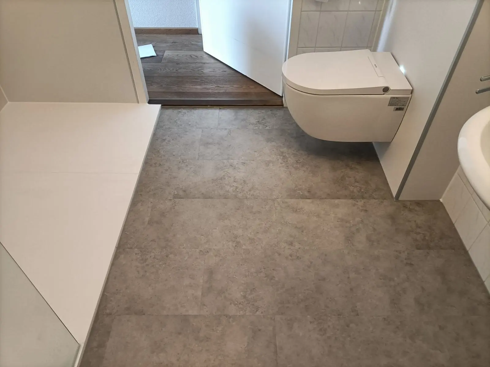 gerenoveerde badkamer met drempelloze inloopdouche, zwevend toilet en grijze tegelvloer. deur naar hal open.